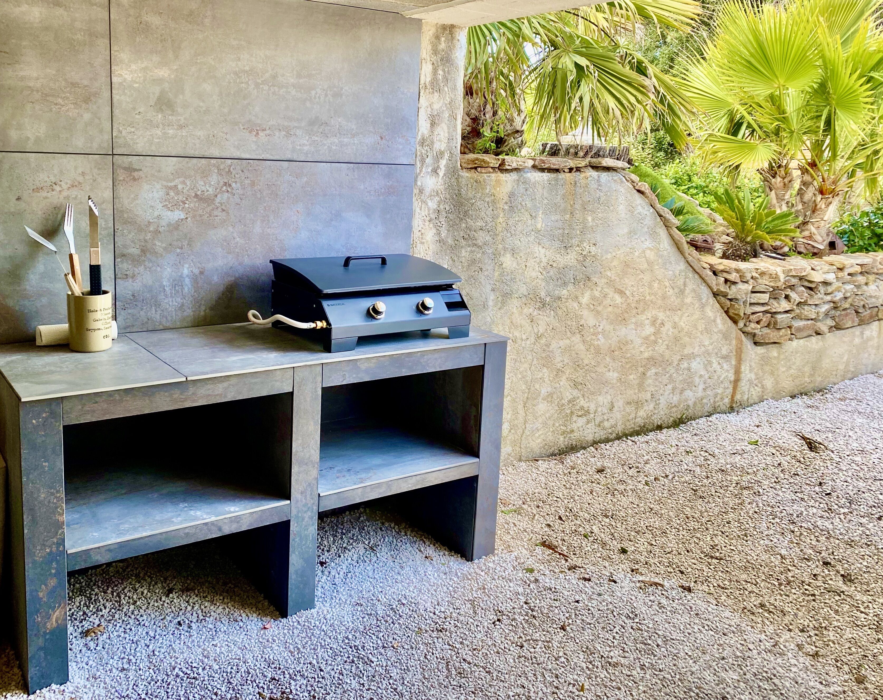 A portable grill on a stone table with a pebble ground, a utensil holder, and a stone wall with greenery.