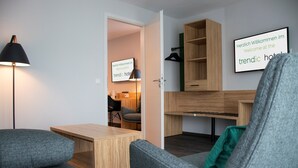 Una habitación de hotel moderna con un sofá gris, un escritorio de madera y una lámpara. Hay un cartel dando la bienvenida a los huéspedes al hotel moderno.