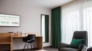 Una habitación de hotel moderna con un escritorio de madera, una silla negra, un sillón gris con un cojín verde y un ventanal con cortinas verdes.
