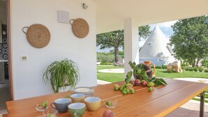 Un comedor con una mesa de madera dispuesta con platos, tazones y vasos, una planta en maceta, y vistas a un jardín y un edificio con techo cónico.