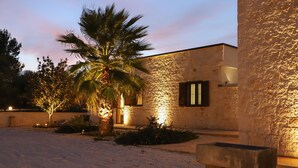 Un edificio de piedra con una palmera en primer plano, un banco de concreto y una entrada bien iluminada.