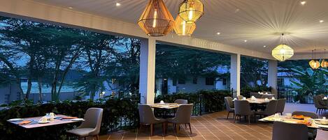 An outdoor dining area with tables and chairs, large windows, and hanging pendant lights.
