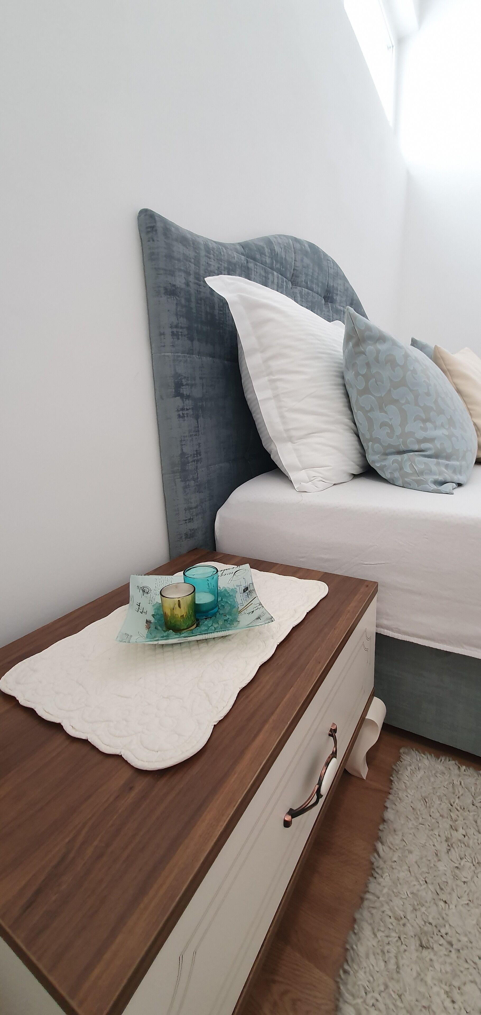 A bedroom with a wooden nightstand, a bed with grey and white bedding, and a white rug.