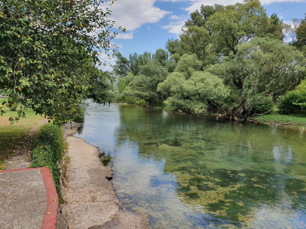 A river with clear water surrounded by trees and a concrete path.