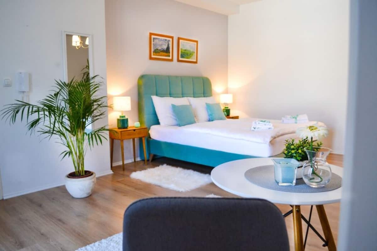 Une chambre moderne avec une tête de lit de couleur turquoise, une grande plante en pot, un tapis blanc et une table ronde agrémentée d’un vase et d’une lampe.