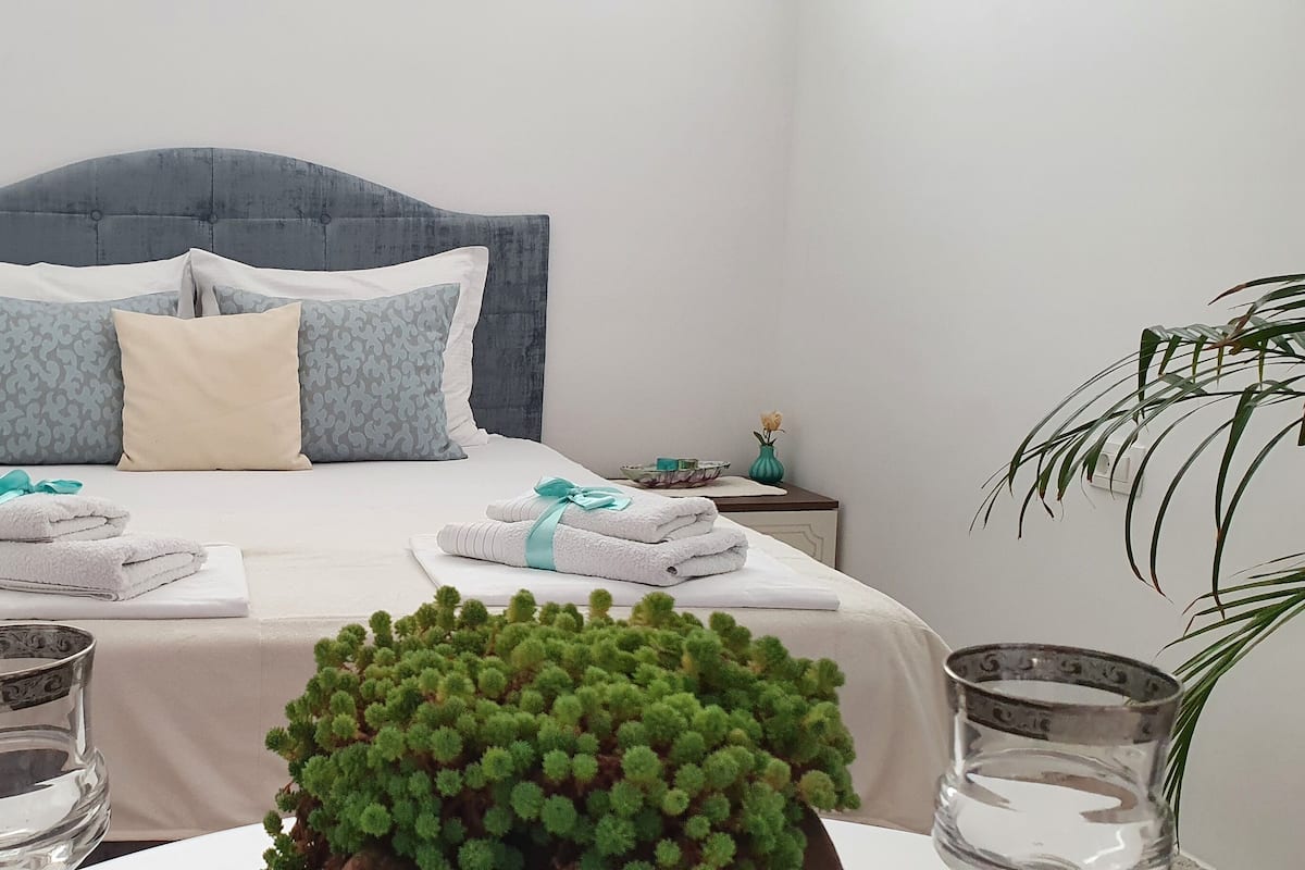 Un lit bien fait, avec une tête de lit grise, deux oreillers blancs et un oreiller à motifs bleus. Deux ensembles de serviettes, ornées de rubans turquoise, sont posés sur le lit. Une petite plante en pot se trouve au premier plan, et un vase en verre avec une fleur jaune est posé sur la table de chevet.