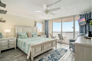 Une chambre à coucher comprenant un lit, des tables de chevet, un bureau, une chaise, un ventilateur de plafond et une vue sur la plage, accessible par une porte coulissante.