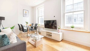 A modern living room with a sofa, coffee table, TV, and dining area. - Fitzrovia Lovely 1BR Flat (London)