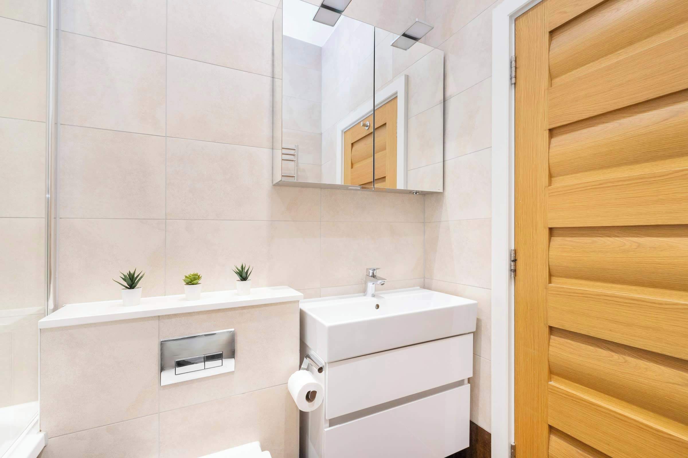 A modern bathroom with a white sink, mirror, and toilet. There are small plants on the countertop and a wooden door.