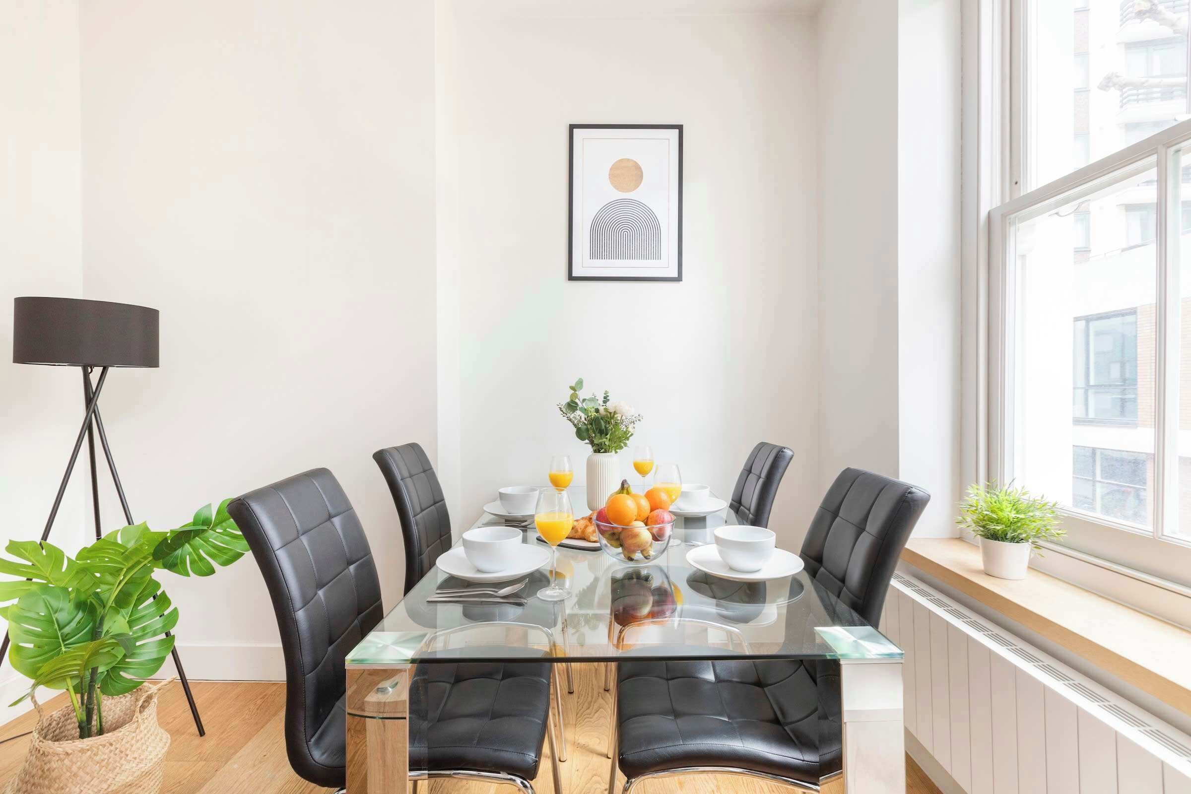 A modern dining area with a glass table, black leather chairs, and a fruit bowl.