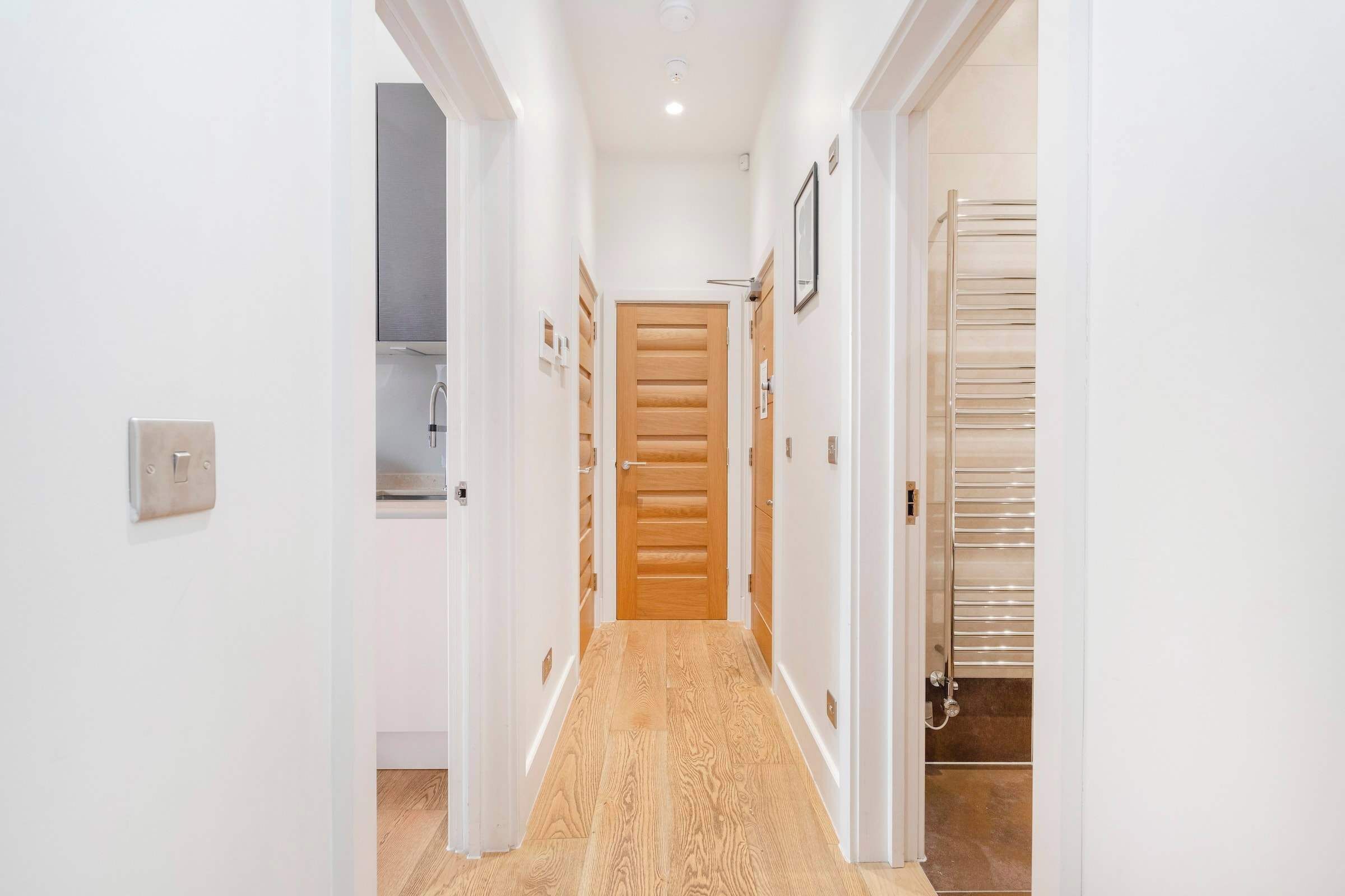 A hallway with a wooden door at the end, white walls, and a tiled floor.