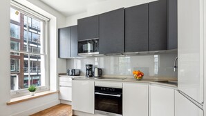 Una cocina moderna con armarios blancos, microondas integrado y lavavajillas. Hay una ventana con una planta en el alféizar y un cuenco de fruta en la encimera.