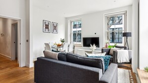 Una sala de estar moderna con un sofá gris, un comedor con mesa y sillas, y ventanas grandes que ofrecen luz natural.