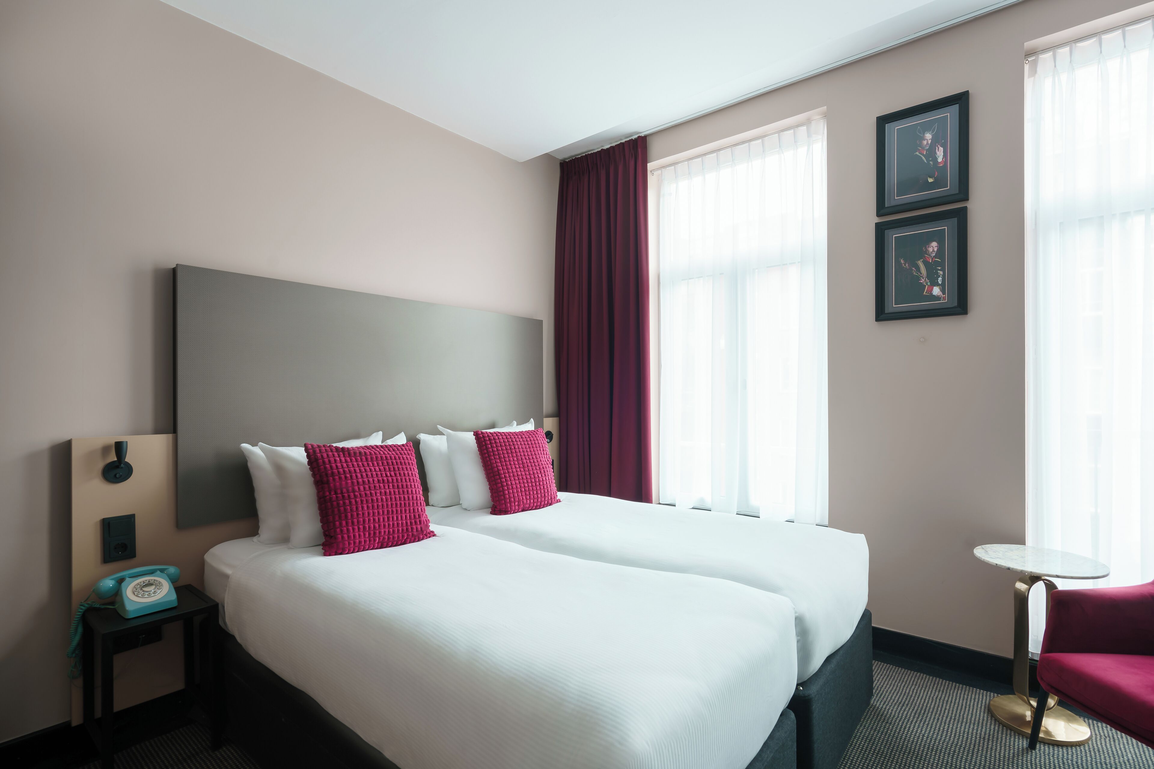 A hotel room with two beds, a small table, a chair, a telephone, and artwork on the wall.