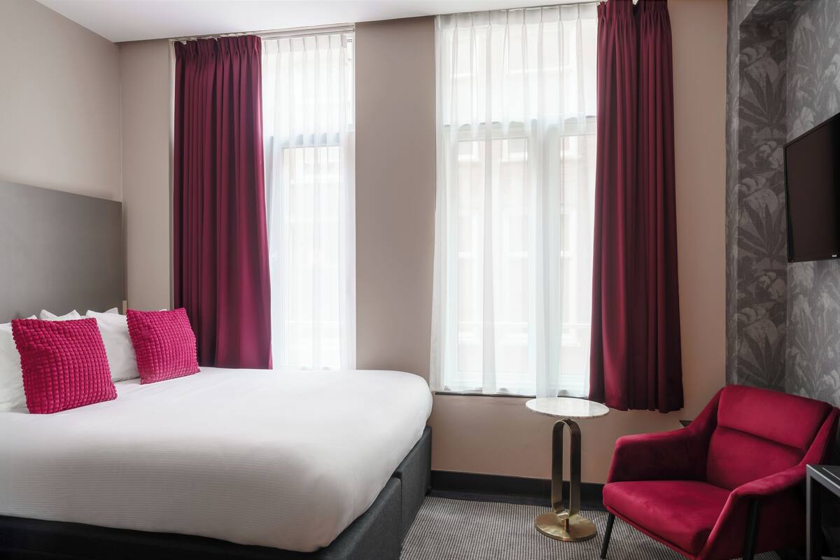 Habitación de hotel con una cama grande, dos almohadas rojas, un sillón rojo, una mesita redonda y un televisor en la pared.