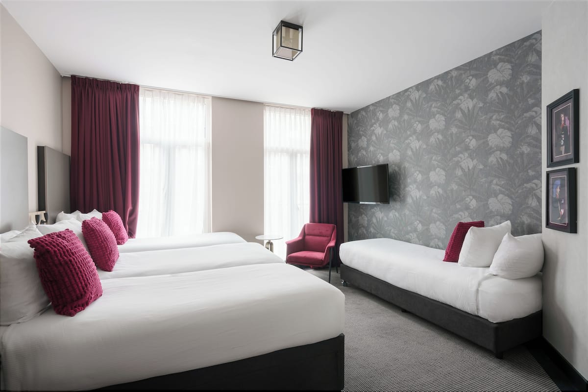 Habitación de hotel con dos camas, un sofá, un sillón rojo, un televisor de pantalla plana y un televisor montado en la pared.