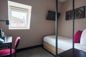 Ein kleiner, moderner Raum mit einem Bett, einem Schreibtisch mit Stuhl, einem Fenster und zwei gerahmten Bildern an der Wand.