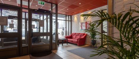 Un hall de hotel con techo rojo, puerta de vidrio de entrada, sofá rojo, una mesita y una planta en maceta.
