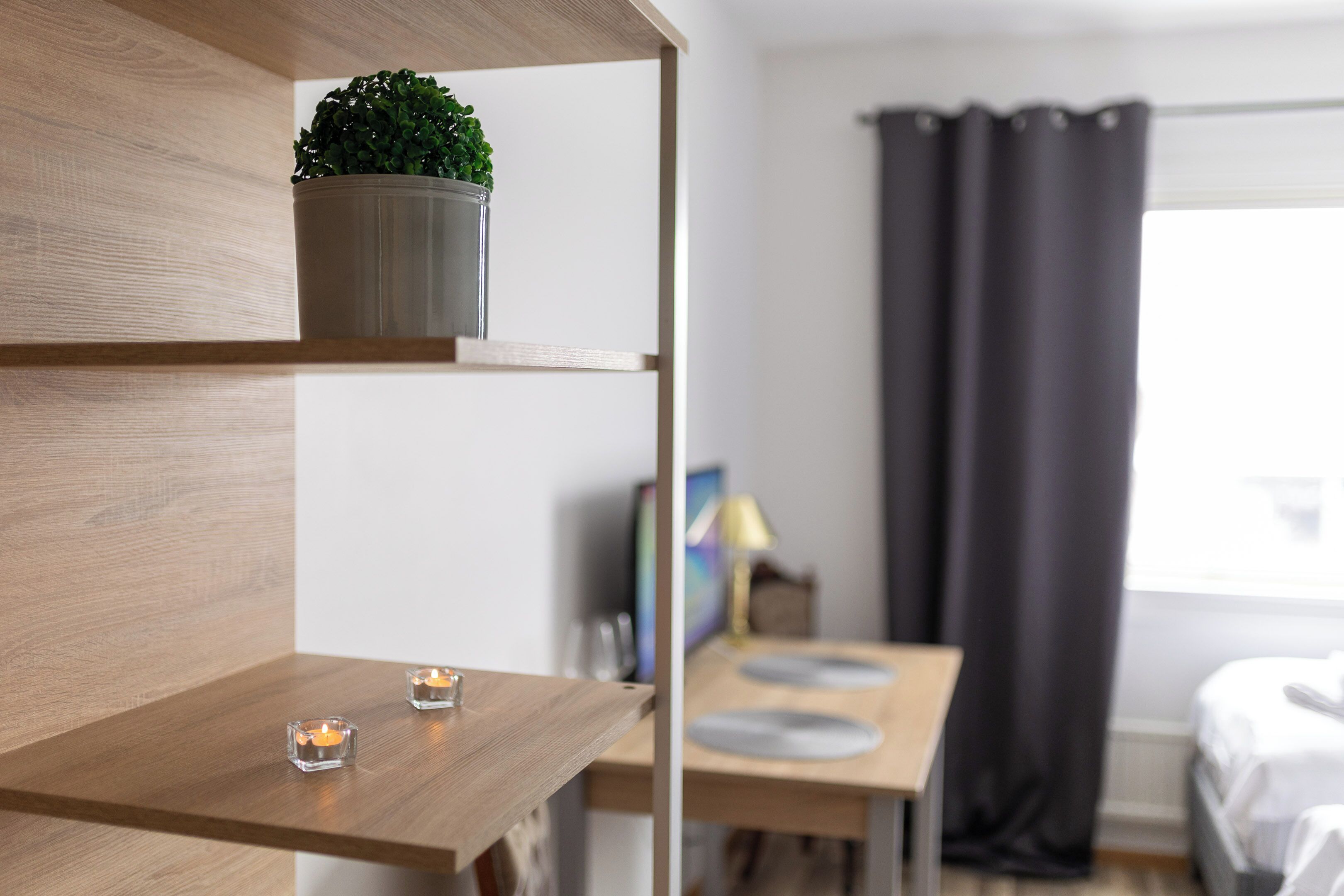 A modern hotel room with a wooden shelf, a potted plant, a small table with candles, a bed, and a window with curtains.