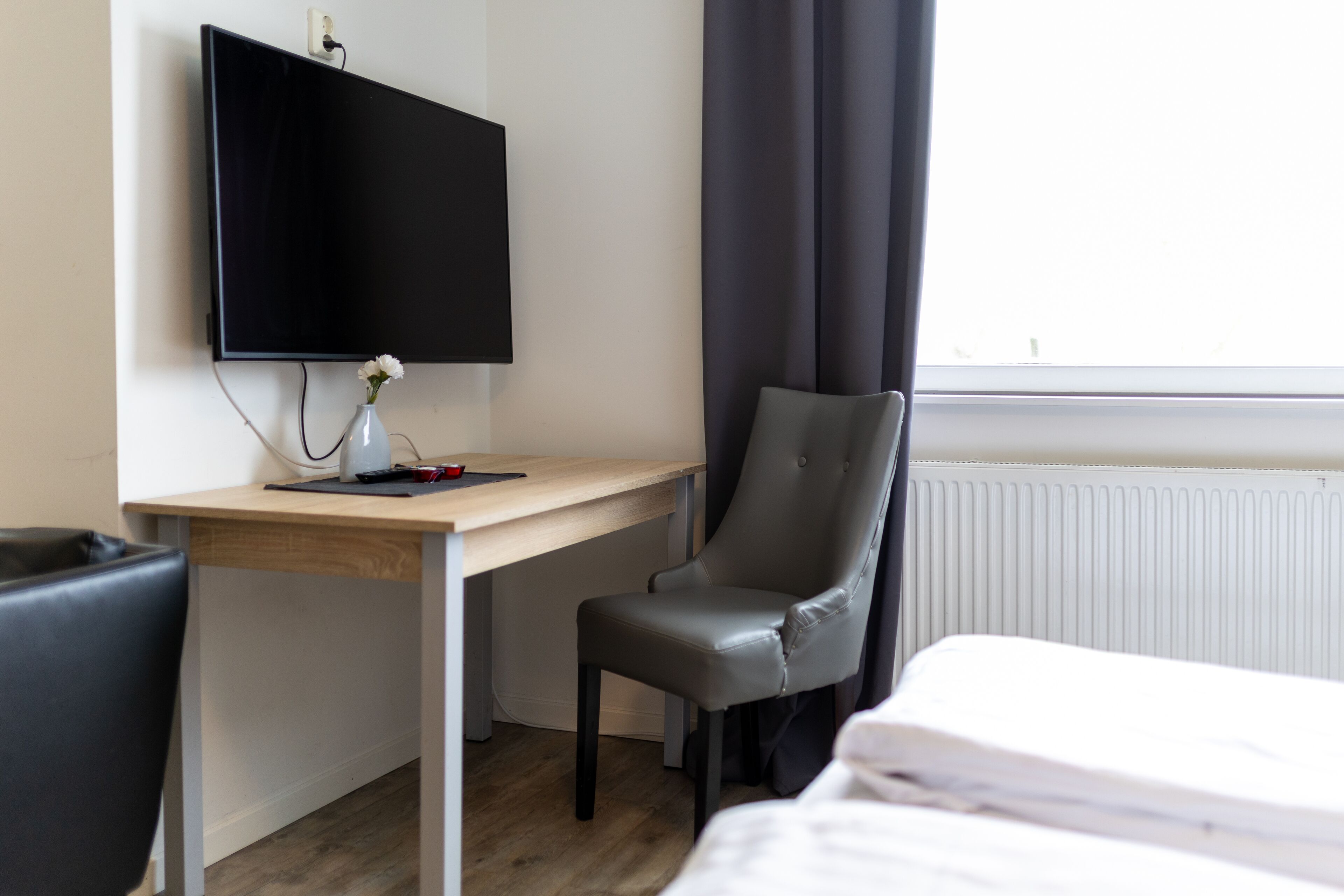 A hotel room with a bed, a desk with a flat-screen TV, a chair, and a window with curtains.