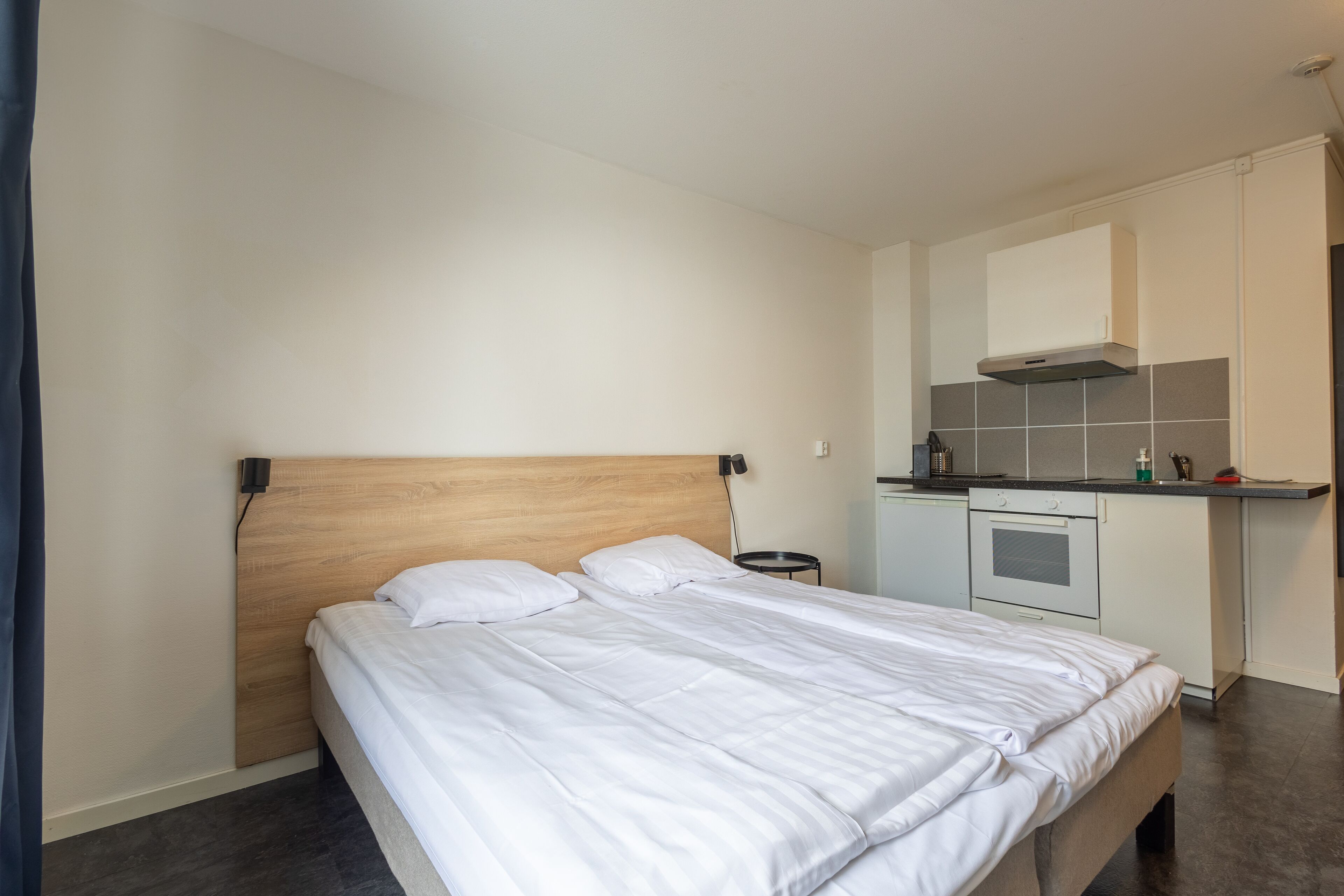 A bedroom with a wooden headboard, a bed with white bedding, and a kitchenette area with a microwave, oven, and sink.