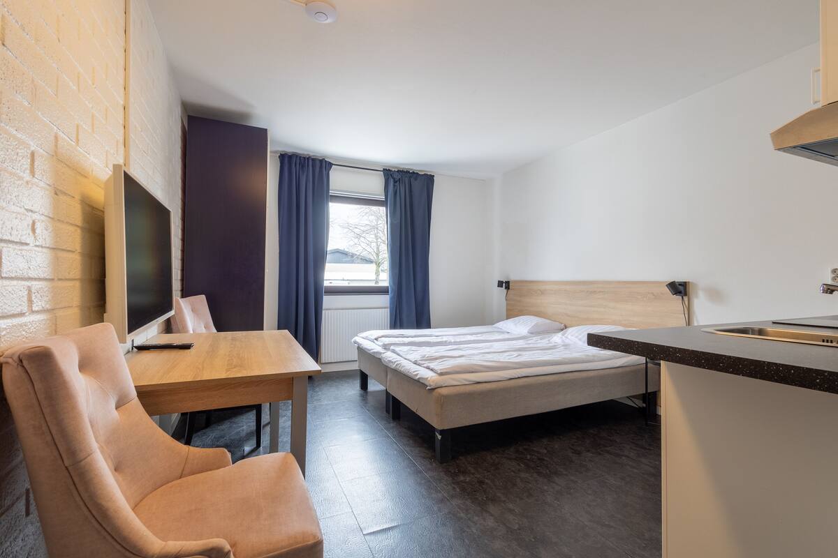 Ein kleines Hotelzimmer mit Bett, Schreibtisch, Stuhl, Fernseher und einer Küchenzeile.