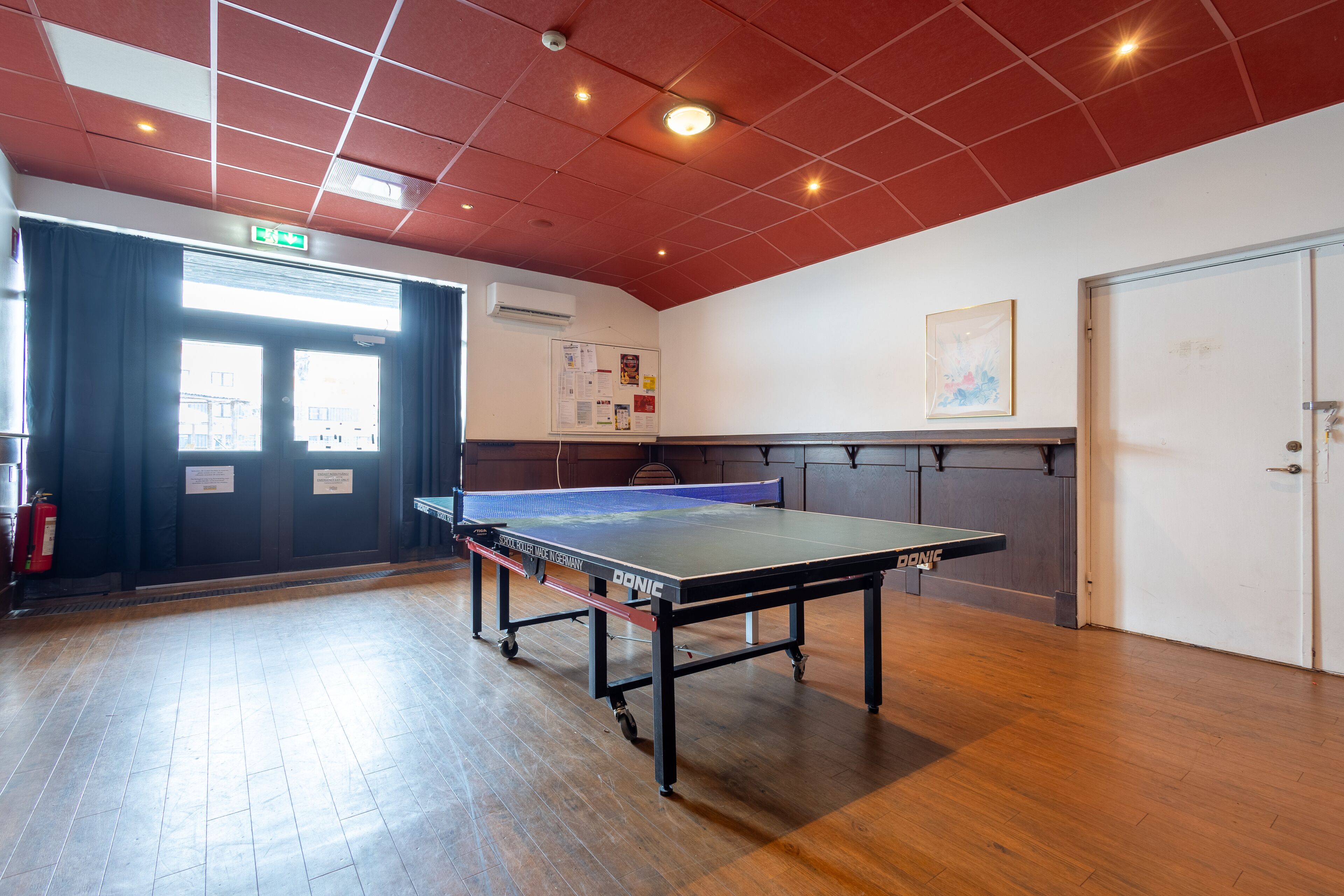 Un tavolo da ping pong in una stanza con pavimento in legno, un estintore e una porta con segnale di uscita.
