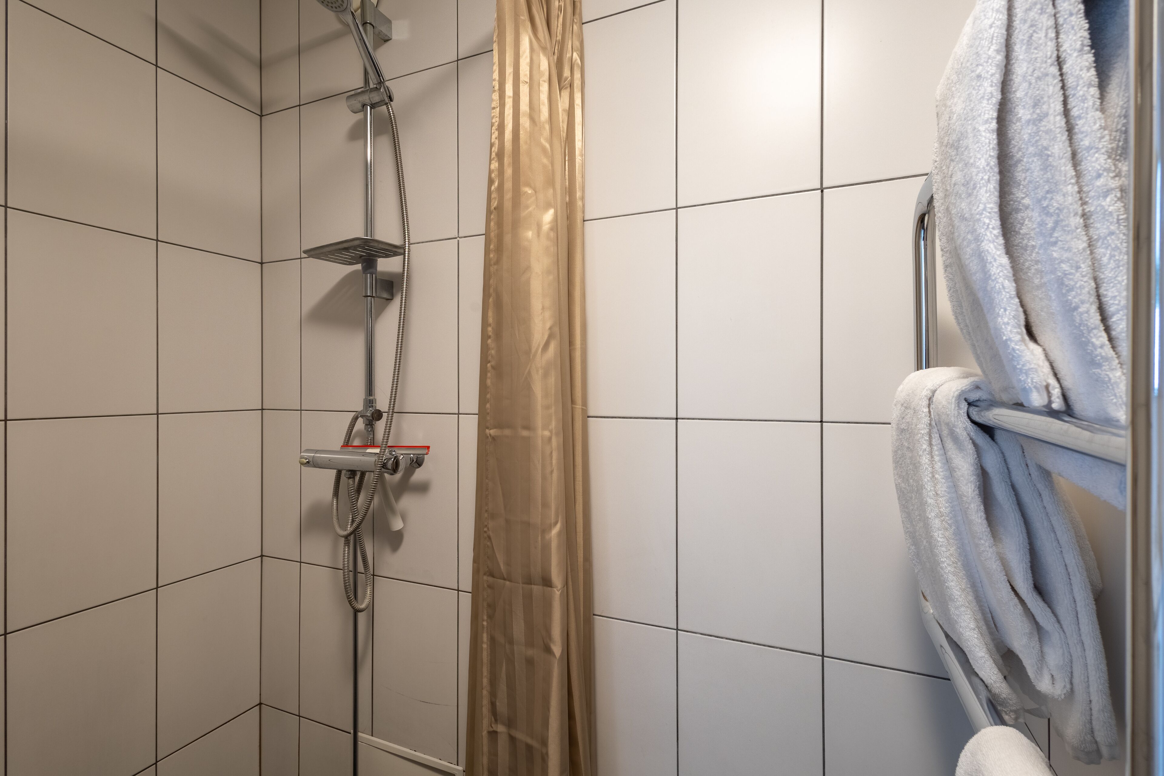 A shower with a handheld showerhead and a vertical rain showerhead, a towel rack with a towel, and a shower curtain.