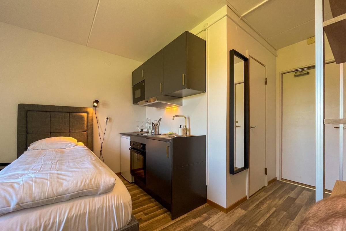 A compact hotel room with a bed, kitchenette, and a door leading to a bathroom.