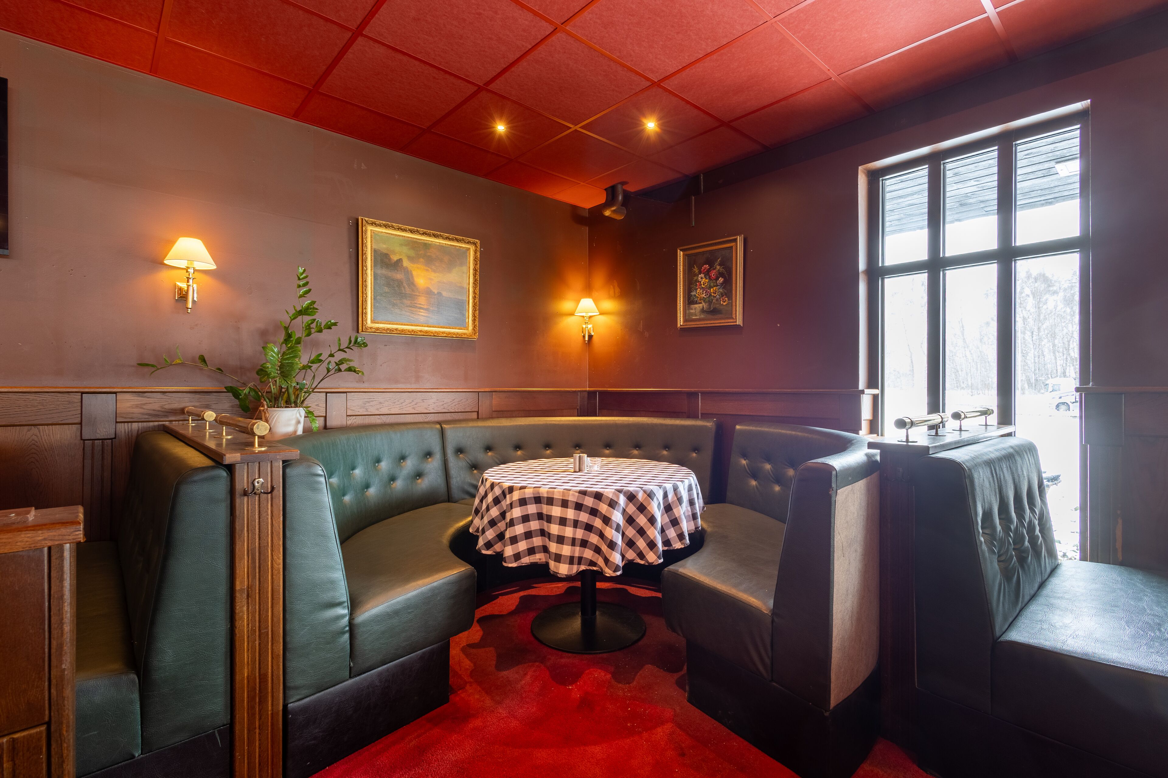 Een restaurantinterieur met een zithoek met ruimtes, een ronde tafel met geruit tafelkleed, wandlampen, kunstwerken in lijsten en een raam met natuurlijk licht.