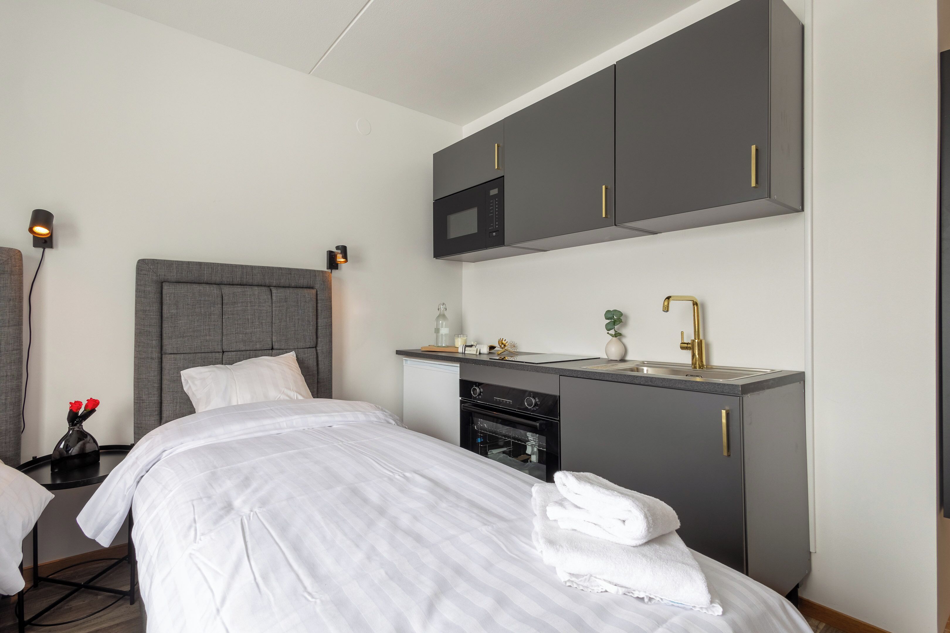 A hotel room with a bed, a built-in desk with a microwave, a sink, and a small oven.