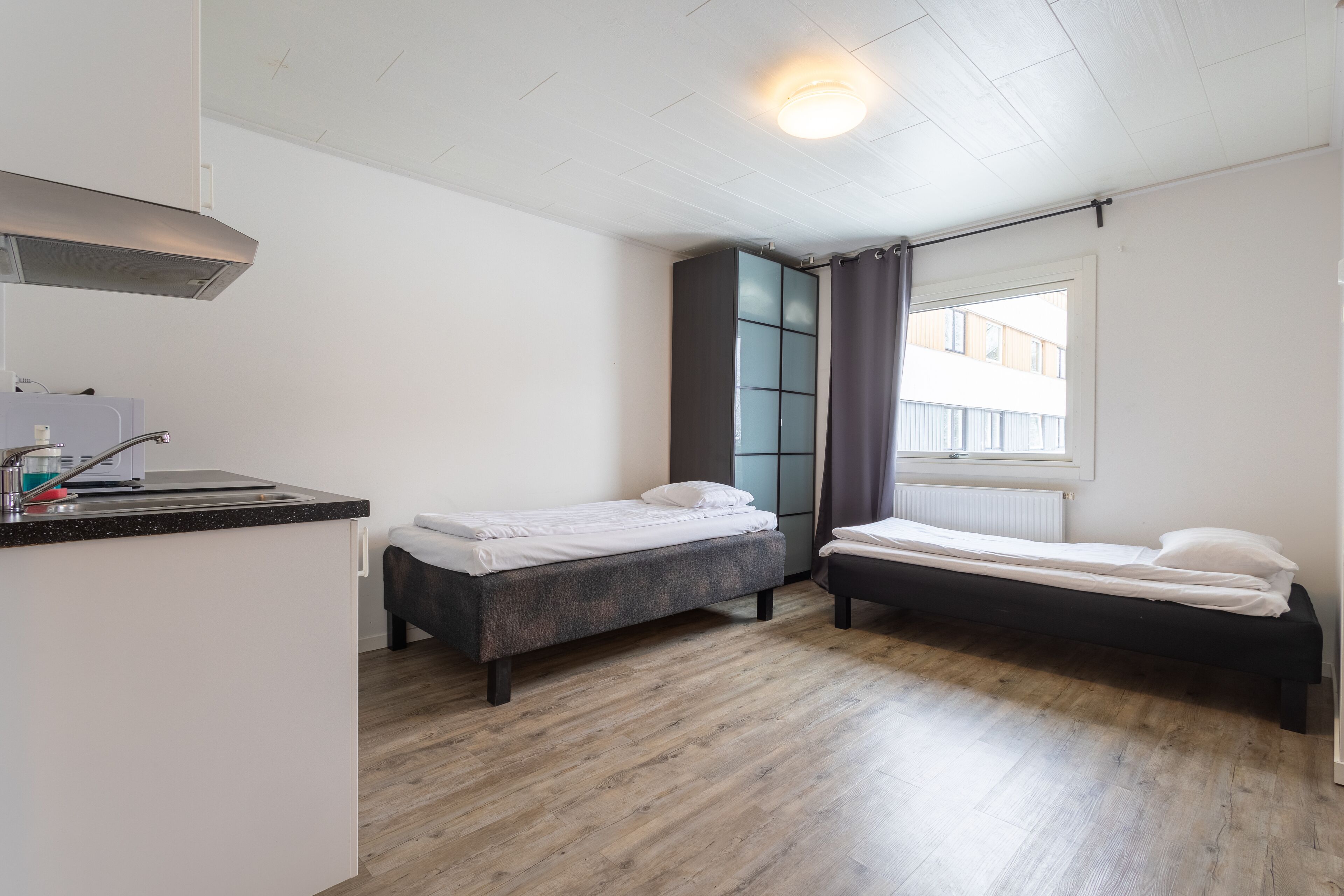 Habitación compacta con kitchenette, dos camas individuales, ventana con cortinas y vista a un edificio.