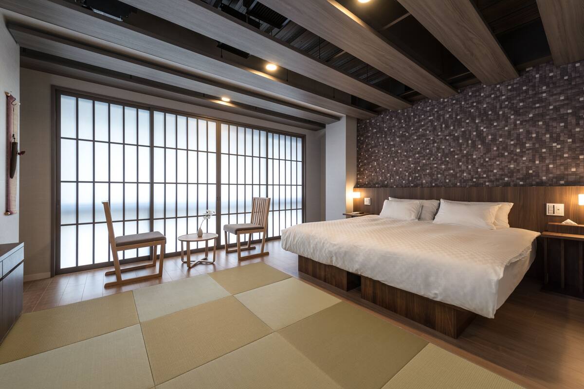 A modern bedroom with a large bed, wooden flooring, tatami mats, a large window with a view, and a textured wall.
