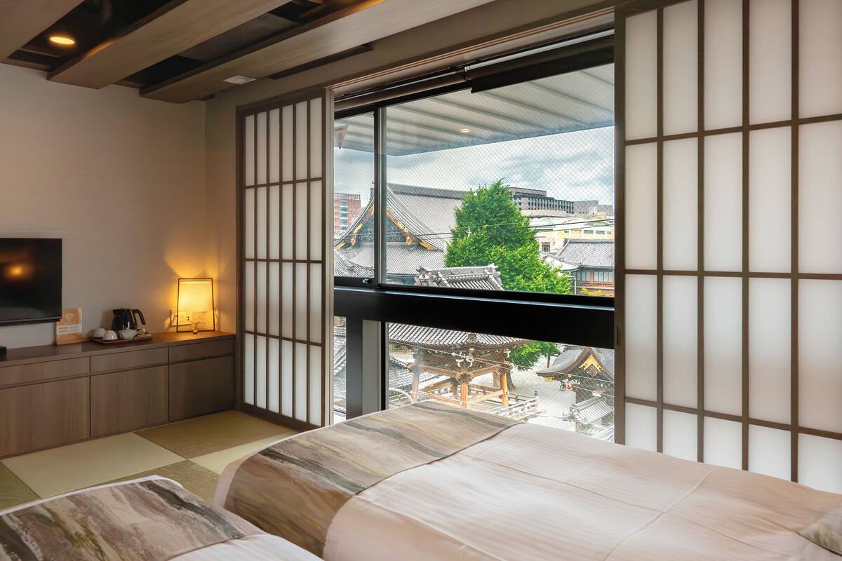 A traditional Japanese-style room with tatami flooring, a bed with a low headboard, a television on a wooden stand, and a view of traditional buildings through a large window.