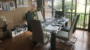 A dining area with a glass table set for four, surrounded by chairs. There are framed pictures on the wall, a clock, and a potted plant. - A house with a rural garden (魚津市)