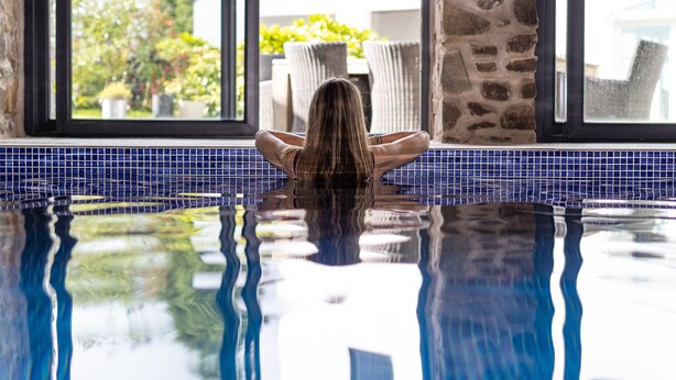 Eine Person sitzt am Pool mit Blick auf Gartenmöbel und Grünpflanzen durch ein Fenster.