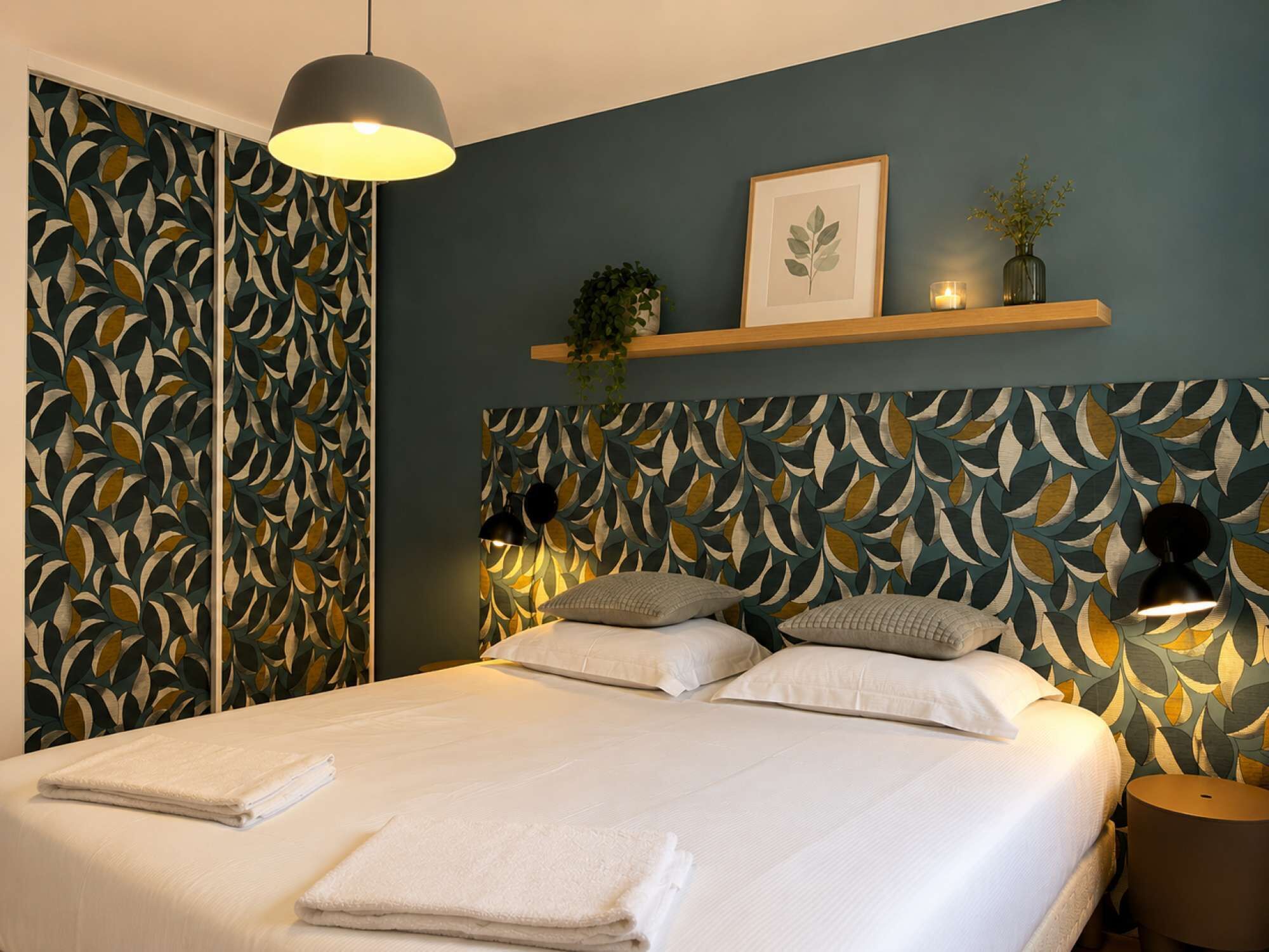 A bedroom with a bed, pillows, a bedside lamp, a nightstand, a shelf with a plant, and a framed picture.