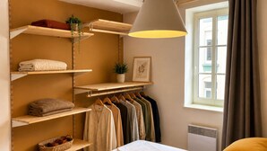 A bedroom with a bed, a hanging light fixture, a window with curtains, and a closet with shelves and hanging clothes. - Le Central (Wimereux)