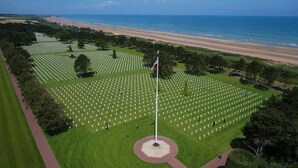 An aerial view of a cemetery with rows of graves, a flagpole, and a beach in the background. - Les 5 rûches (Courseulles-sur-Mer)