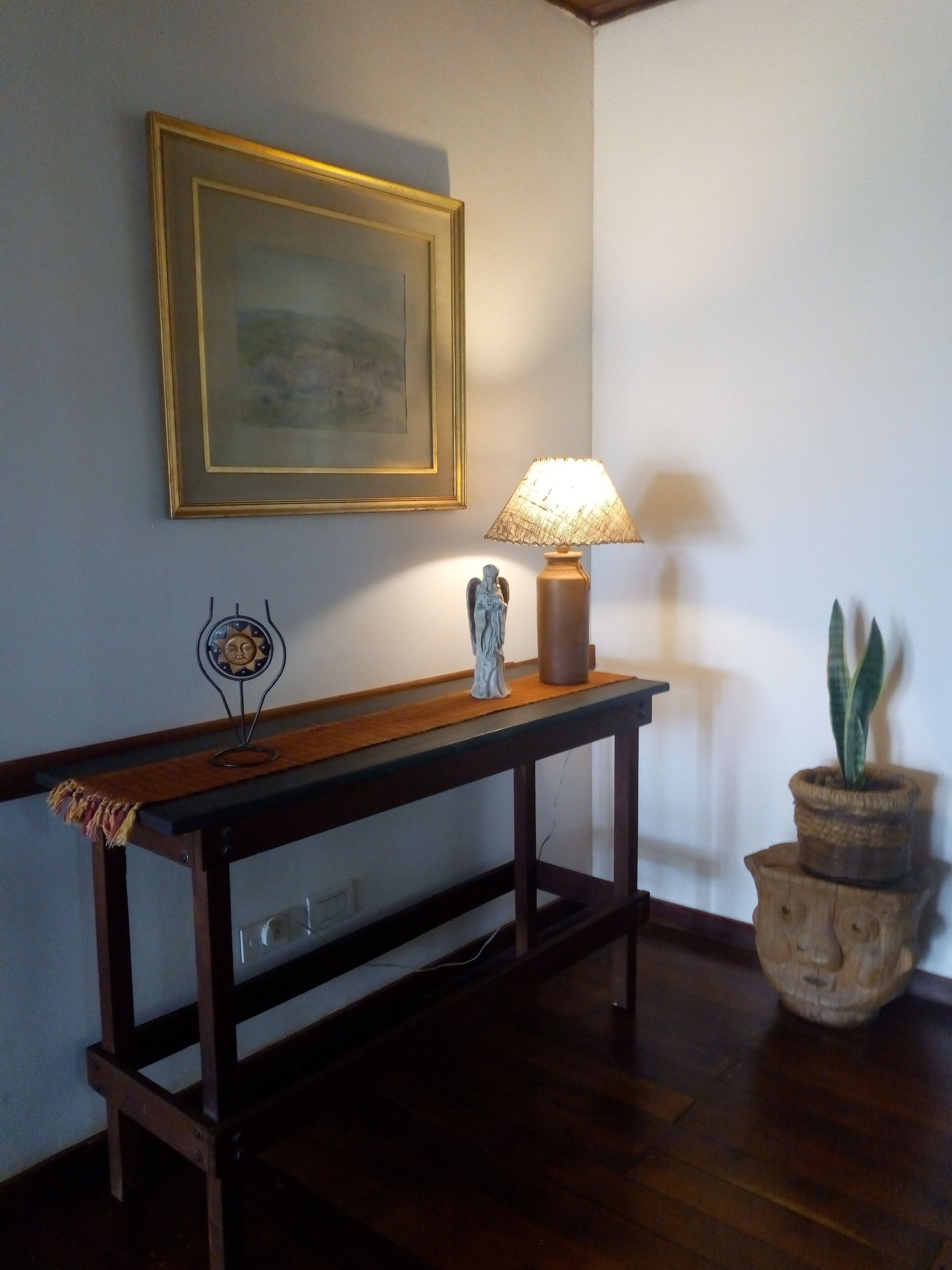 A wooden console table with a lamp and a small statue, a framed picture on the wall, and a potted plant.
