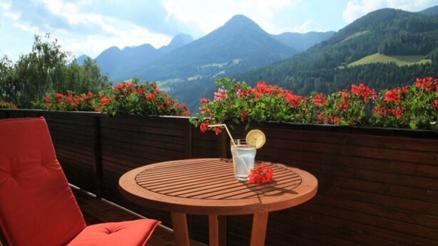 A round wooden table with a single chair, a glass of water with a lemon slice, and red flowers on a balcony with a mountain view.