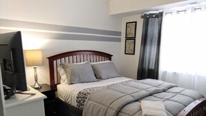A neatly made bed with a grey comforter and pillows, a bedside lamp, a television on a stand, and a window with curtains. - Awesome Condo in Central Raleigh (Raleigh)
