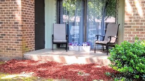 A covered porch with two chairs, a potted plant with flowers, and a brick wall. - Awesome Condo in Central Raleigh (Raleigh)