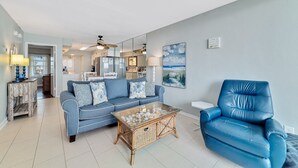 Uma sala de estar com um sofá azul, uma poltrona azul, uma mesa de centro de vidro e uma área de jantar ao fundo.