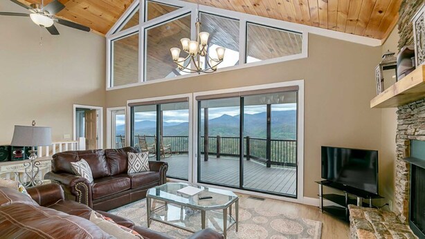 A spacious living room with a large sofa, a coffee table, and a TV. There are large windows offering a view of mountains.