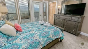A bedroom with a bed, dresser, television, and a view through the window blinds.
