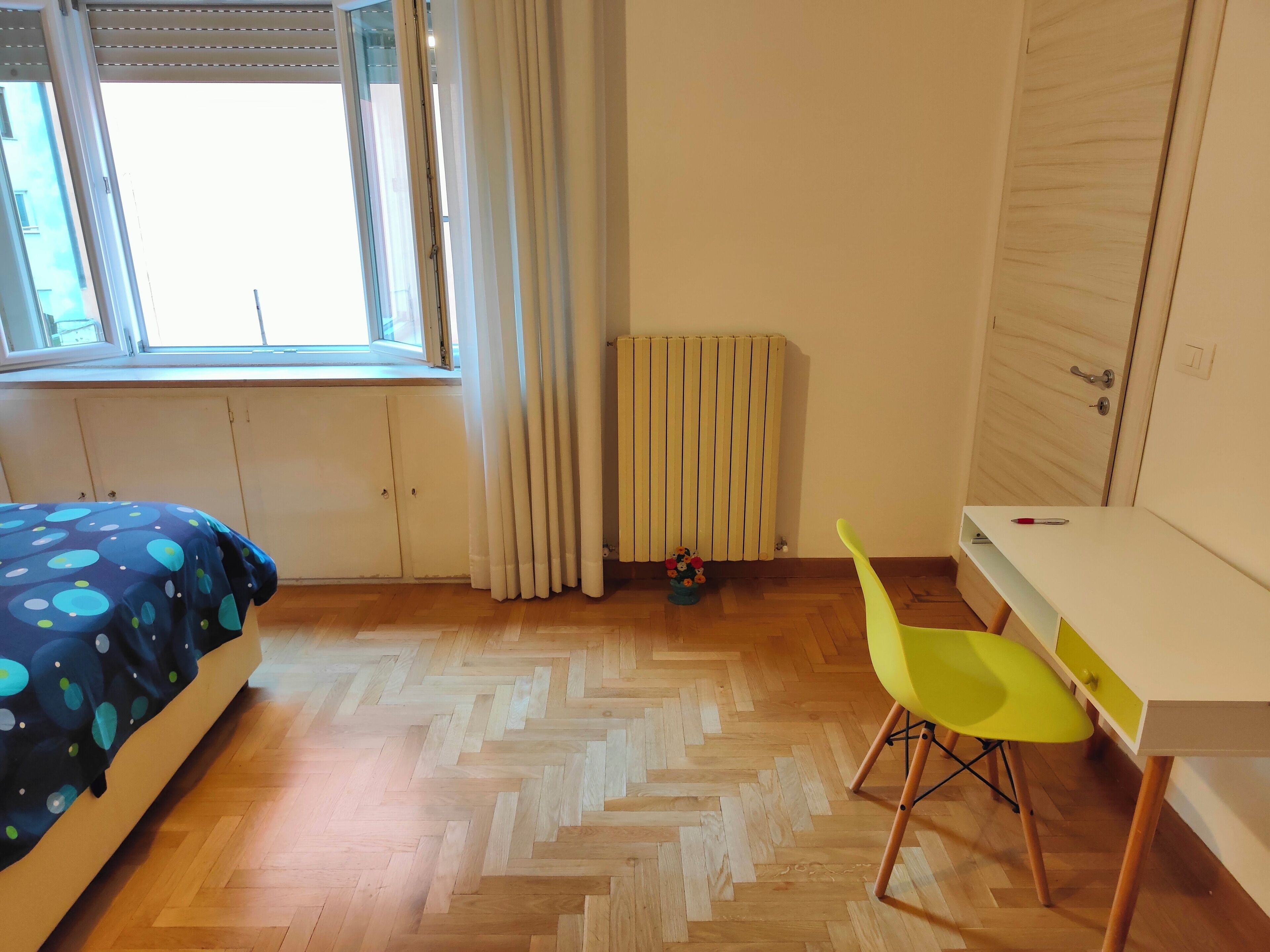 A bedroom with a bed, a desk, a chair, a radiator, and a window.