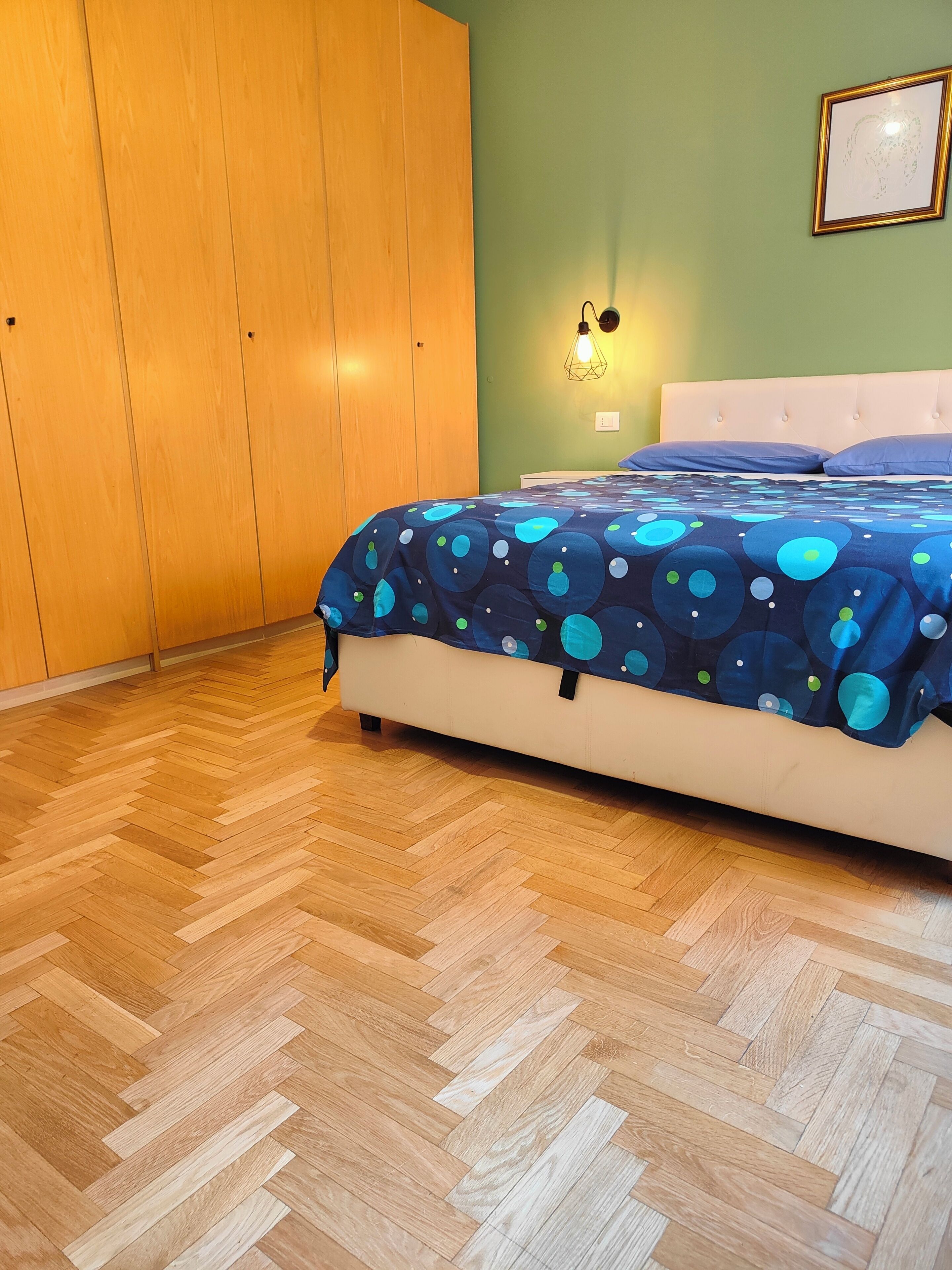 A bedroom with a wooden floor, a bed with a blue patterned bedspread, a white headboard, and a wall-mounted light fixture.
