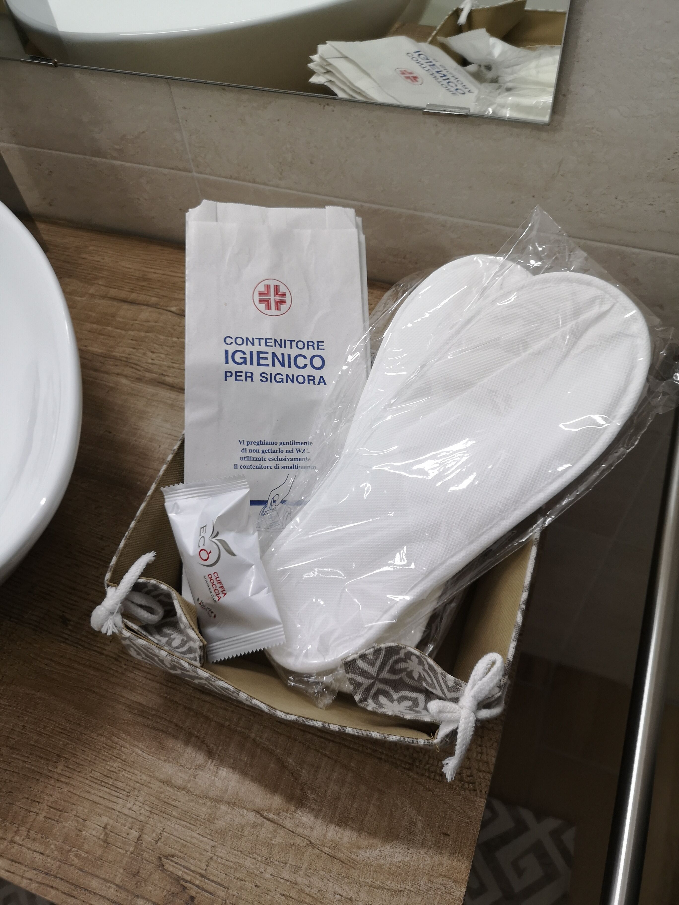 Un baño con lavamanos, espejo y una caja con una toalla blanca y un pequeño kit de artículos de aseo.