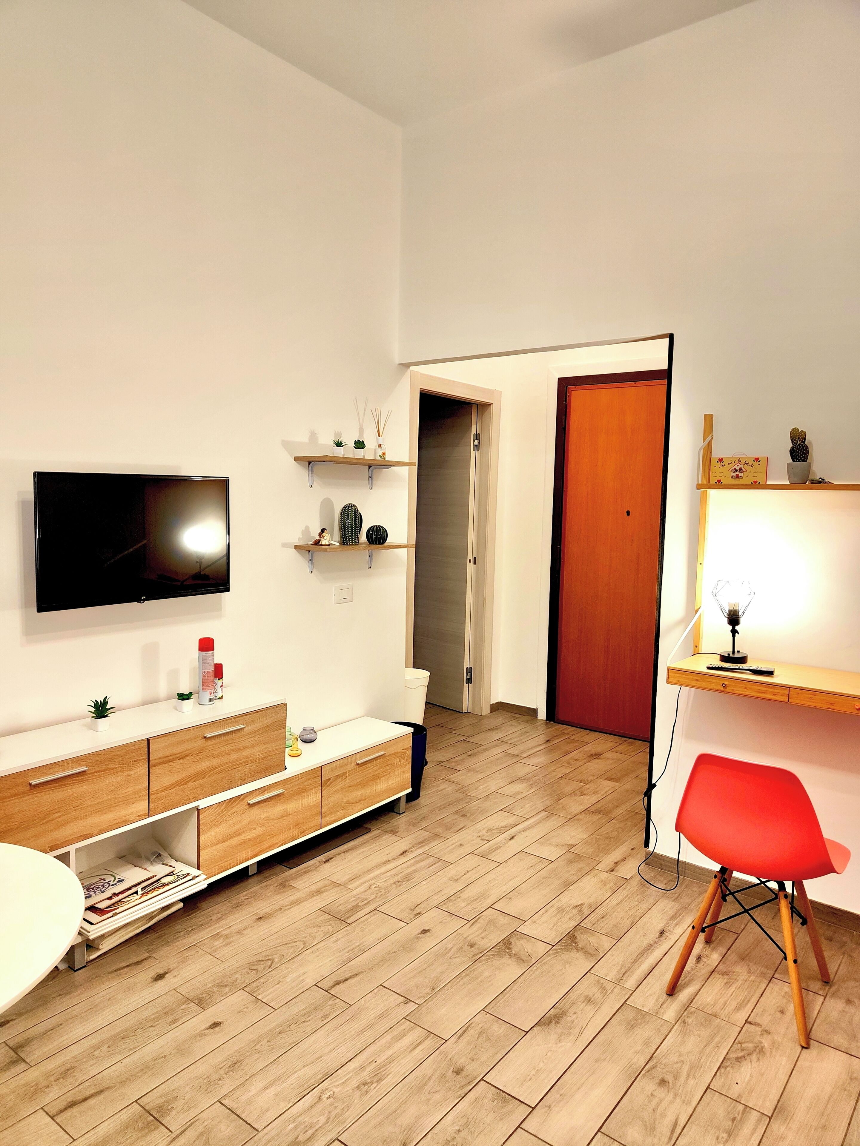 A modern living room with a flat-screen TV, wooden flooring, a red chair, and a wooden desk with a lamp.